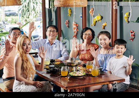 Felice grande famiglia vietnamita seduti al tavolo per la cena con il tradizionale cibo asiatico, salutando con le mani e guardando la fotocamera Foto Stock
