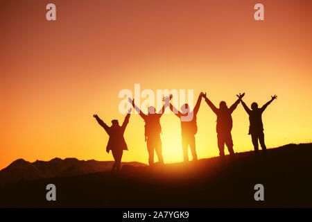 Gruppo di felice popoli sagome sta con le braccia sollevate contro il tramonto e le montagne Foto Stock