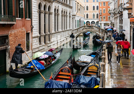 Giorno di pioggia a Venezia con gondolieri turisti a remi lungo i canali accanto ai camminatori con ombrelloni - Venezia Italia Foto Stock