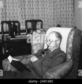 Anni '1950, storico, un uomo anziano in abito e cravatta, seduto su una poltrona fumando una pipa e leggendo un libro, Inghilterra, Regno Unito. Foto Stock