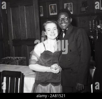 Anni '1950, storica, in posa per una foto a un ballo nero, una giovane donna inglese con un giovane africano, Inghilterra, Regno Unito. La Gran Bretagna del dopoguerra vide molte persone provenienti dai paesi del Commonwealth arrivare in Gran Bretagna per lavorare e studiare e si tennero scambi culturali e danze per introdurli allo stile di vita britannico. Foto Stock