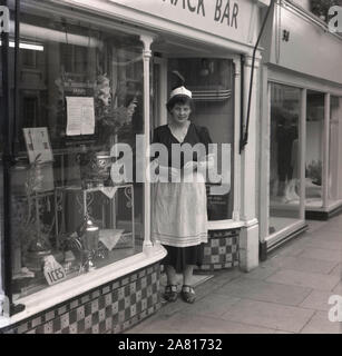 Anni '1950, una cameriera in uniforme in piedi per la sua foto all'ingresso di una sala da tè o snack bar dell'epoca, Inghilterra, Regno Unito. Foto Stock