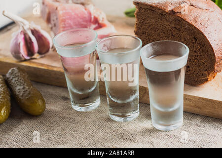 Tre Bicchieri di vodka sul tavolo sullo sfondo di snack - lardo, aglio, prezzemolo, pane, cetriolo Foto Stock