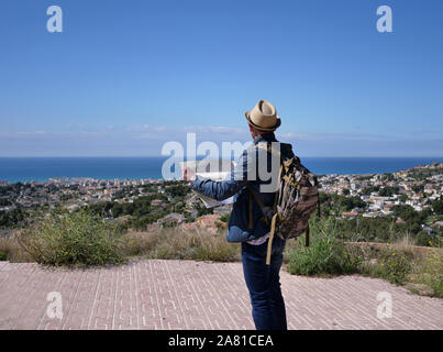 Un giovane uomo con una mappa nelle sue mani sulla montagna guarda al villaggio sotto la montagna, vista posteriore Foto Stock