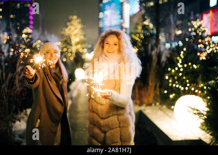 Foto di due donne con luci di bengala sulla passeggiata invernale sullo sfondo di abete decorato in sera in città Foto Stock