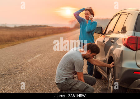 Giovane riparazione auto pneumatico sgonfio sulla strada in autunno il tramonto e la chiamata per assistenza stradale, il fuoco selettivo Foto Stock