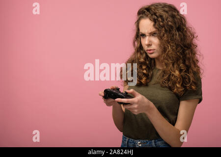 Piuttosto gamer ragazza con i capelli ricci azienda controller di gioco e concentrati durante la riproduzione di un videogioco isolato su sfondo rosa in studio Foto Stock