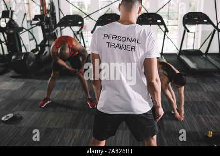 Vista posteriore del personal trainer di supervisione atleti multiculturale stretching in palestra Foto Stock