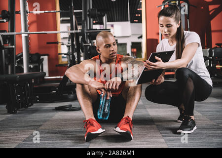 Attraente trainer mostra appunti per african american sportsman seduto sul pavimento Foto Stock