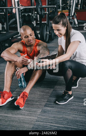 Trainer sorridente mostra appunti per allegro african american sportsman seduto sul pavimento Foto Stock