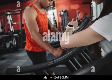 Vista ritagliata del trainer guardando il tracker di fitness mentre in piedi nelle vicinanze African American sportsman in esecuzione sul tapis roulant Foto Stock