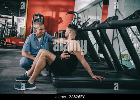 Giovane atleta guardando sorridente americano africano medico mentre soffrono di dolore addominale Foto Stock