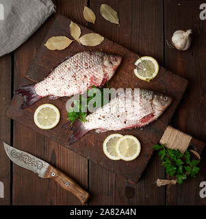 Fresco pesce carassio cosparso con spezie e le fette di limone e giace su di un marrone tagliere in legno, tavolo in legno da schede, vista dall'alto Foto Stock