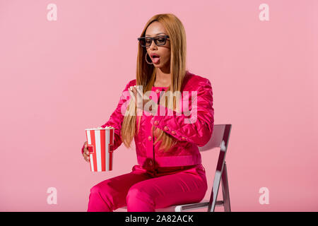 Attraente american donna seduta su una sedia e mangiare popcorn isolati su rosa, moda bambola concept Foto Stock