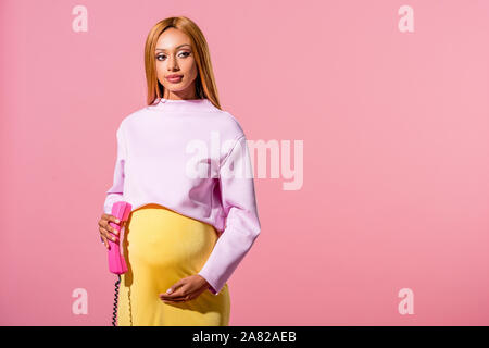 Attraente, africana incinta donna americana tenendo il ricevitore isolati su rosa, moda bambola concept Foto Stock