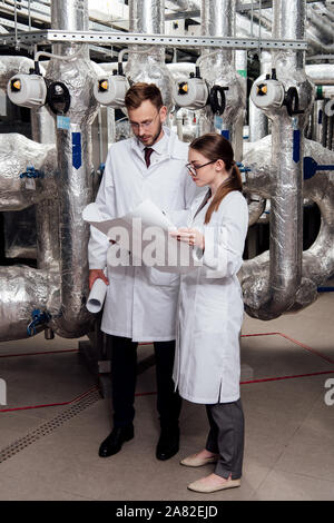 Gli ingegneri in bicchieri e camici guardando blueprint vicino il sistema del compressore d'aria Foto Stock