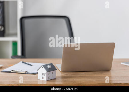 Modello di casa, laptop e appunti con la penna sulla scrivania in legno vicino a sedia ufficio Foto Stock