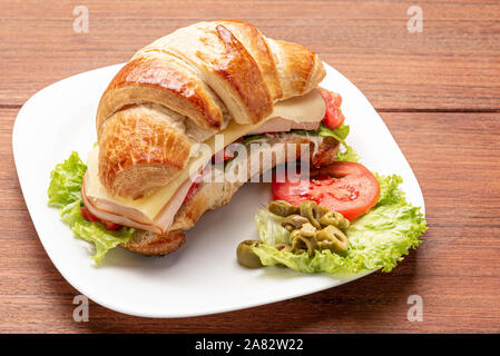 Croissant sandwich con insalata, prosciutto, formaggio, pomodori e olive su sfondo di legno. La colazione del mattino concetto. Sana e fast food. Isolato. Foto Stock