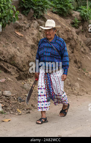 Un vecchio contadino Maya che indossa il vestito tradizionale di San Pedro La Laguna, Guatemala, passeggiate per la sua piccola fattoria trama su una strada sterrata che porta un machete Foto Stock