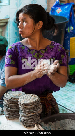 Una giovane donna Maya in abito tradizionale rende blue tortilla di mais nel mercato a Santiago Atitlan, Guatemala, pacche di essi tra le sue mani. Foto Stock