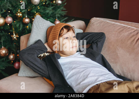 Attraente funny tween ragazzo con i capelli scuri in hat aspettando il nuovo anno il divano a L'albero di Natale sfondo Foto Stock