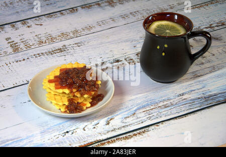 Una ceramica nera tazza di tè con una fettina di limone e una piastra con cialde Viennese e confettura di fragole. Close-up. Foto Stock