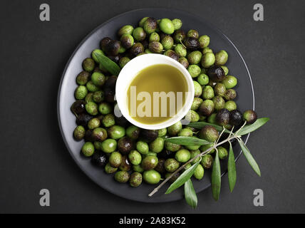 Ciotola di freschi olio extra vergine di oliva e di olive verdi con foglie in una piastra isolata su sfondo scuro. Vista superiore Foto Stock