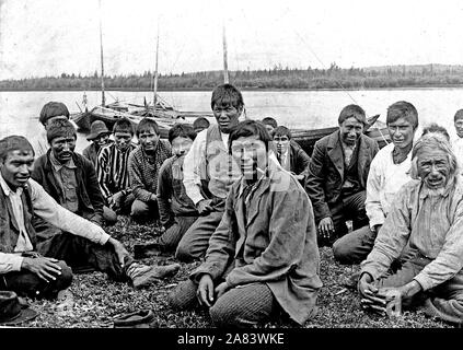 Indian uomini e ragazzi in abito occidentale, seduti lungo il litorale, barche in background. 1900-1923 della Baia di Hudson indiani Foto Stock