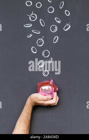 Concetto di risparmio di denaro con una colorazione rosa salvadanaio tenuto in una mano di fronte a una lavagna con disegnati a mano delle monete in euro Foto Stock