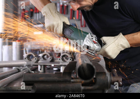 Uomo lavoro in officina di casa garage con smerigliatrice angolare, occhiali di protezione e guanti di costruzione, metallo levigatura fa scintille closeup, fai da te e del concetto di artigianato Foto Stock