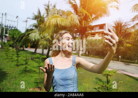 Donna allegra effettuare chiamate video con smart phone, viaggiando verso la soleggiata della Thailandia con palme e prato verde Foto Stock