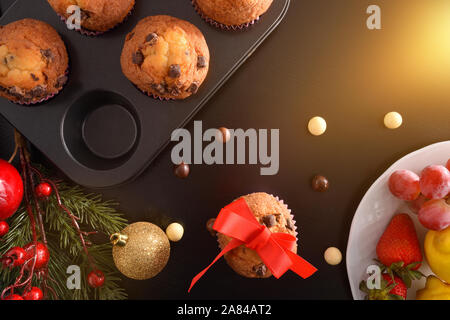 Fresco cotto in casa muffin al cioccolato su stampo sulla tavola nera con decorazione di Natale e un piatto di frutta. Composizione orizzontale. Vista dall'alto. Foto Stock