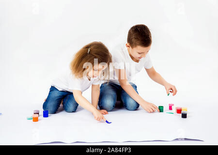 Due belle bambino amici un ragazzo e una ragazza in camice bianco e blu jeans, alla moda capelli stile, scalzi, disegnare immagini sul foglio di carta bianca da parte delle vernici isolato su bianco. Studio shot. Foto Stock