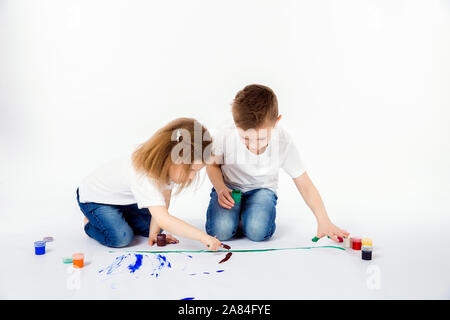 Due belle bambino amici un ragazzo e una ragazza in camice bianco e blu jeans, alla moda capelli stile, scalzi, disegnare immagini sul foglio di carta bianca da parte delle vernici isolato su bianco. Studio shot. Foto Stock