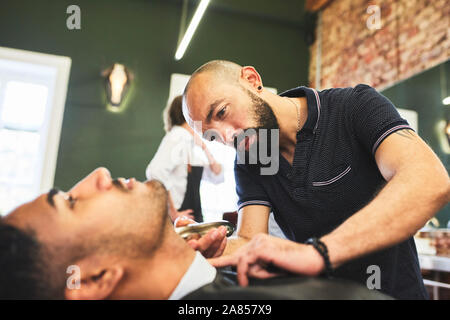 Maschio focalizzato barbiere dando il cliente una rasatura in Barberia Foto Stock