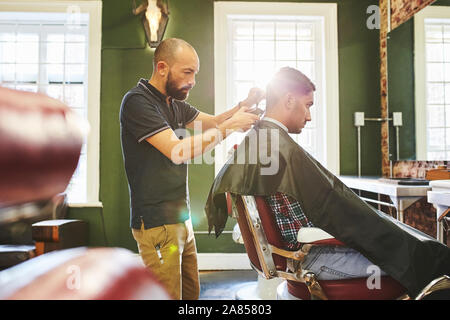 Maschio focalizzato barbiere di dare al cliente un taglio di capelli in Barberia Foto Stock