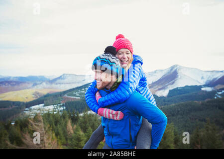 Coppia felice piggybacking con le montagne sullo sfondo, Lake District, REGNO UNITO Foto Stock