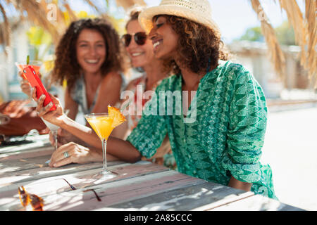 Felici donne amici utilizzando smart phone e bere cocktail a sunny beach bar Foto Stock