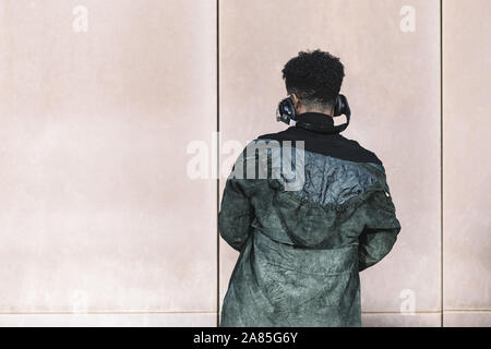 Vista posteriore di un uomo nero con le cuffie per ascoltare la musica di fronte a un muro, tecnologia e concetto di stile di vita Foto Stock