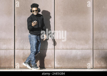 Cool nero africano uomo con casual abbigliamento moderno e ascolto in cuffia la musica del suo telefono sorridendo a piedi nella parte anteriore di una parete, la tecnologia e la vite Foto Stock