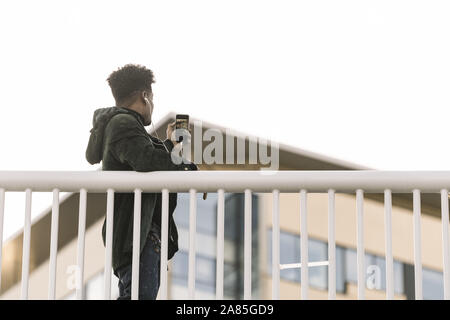 African uomo in nero gli auricolari di scattare una foto a sunny street della città con il suo telefono cellulare, selfie per le reti sociali, tecnologia e stile di vita con Foto Stock