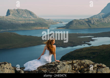 Retro della ragazza in abito da sposa seduta sulle rocce della montagna e guardando al fiordo, escursionismo in colline Foto Stock