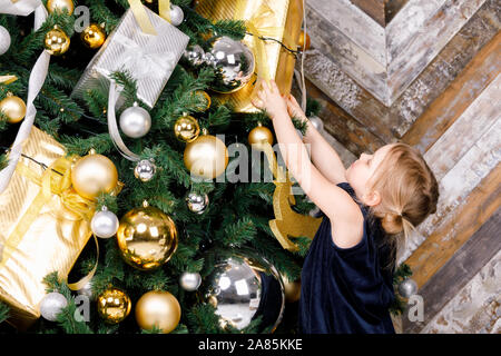 Elementare di età ragazza indossa vestito blu tirando le mani cercando di raggiunge per avvolgere presente appeso su albero di Natale a casa Foto Stock