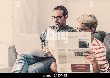 Giovane ragazzo in lettura di un quotidiano mentre il padre sta lavorando con il computer portatile, seduto su un divano a casa. Informazioni con tradizionale rivista cartacea e ultimo Foto Stock