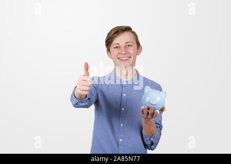 Ragazzo adolescente holding salvadanaio con il pollice fino segni su sfondo bianco. Educazione finanziaria il concetto di risparmio. Foto Stock