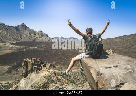 Uomo sportivo braccia aperte godendo un bel giorno dopo la scalata da solo. Ragazzo caucasico con zaino che guarda lontano seduto su una montagna che si affaccia sulla scogliera Foto Stock