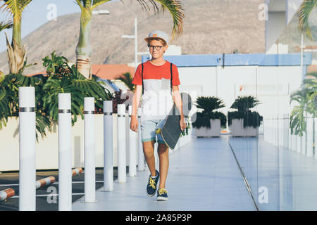 Bambino alla moda a camminare in una città moderna di contesto. Felice ragazzo con lo skateboard in mano e uno zaino cammina da sola. Vista frontale delle persone che passeggiano Foto Stock