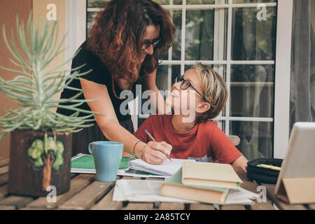 Giovane studente di fare i compiti a casa con i libri di scuola, quotidiano e pad digitale aiutato da sua madre. La mamma la scrittura sul copybook insegnando il suo figlio. Foto Stock