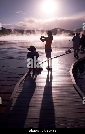 I visitatori si stagliano contro il sole e la crescita del vapore a Grand Prismatic Hot Springs nel Parco Nazionale di Yellowstone, Wyoming USA Foto Stock