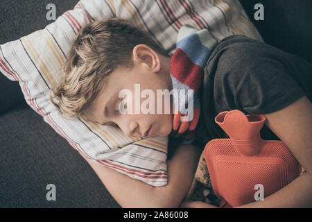 Ragazzo malato a letto con coperta di lana e bottiglie di acqua calda. Triste teen con l'influenza si appoggia a casa da solo in una fredda giornata invernale. Bambino con infettare stagionali Foto Stock